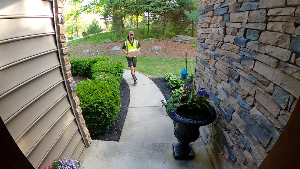 A person in a high-visibility vest carries a clipboard, walking on a residential path lined with green plants and decorative stone walls, suggesting a professional visit.