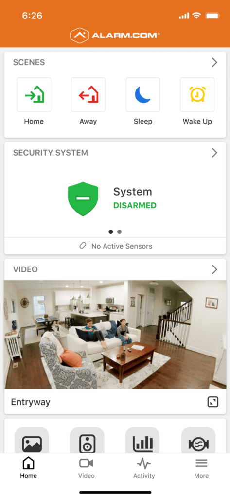The image shows a smartphone screen with the Alarm.com app open, displaying home automation options and a video feed of two people sitting in a living room.
