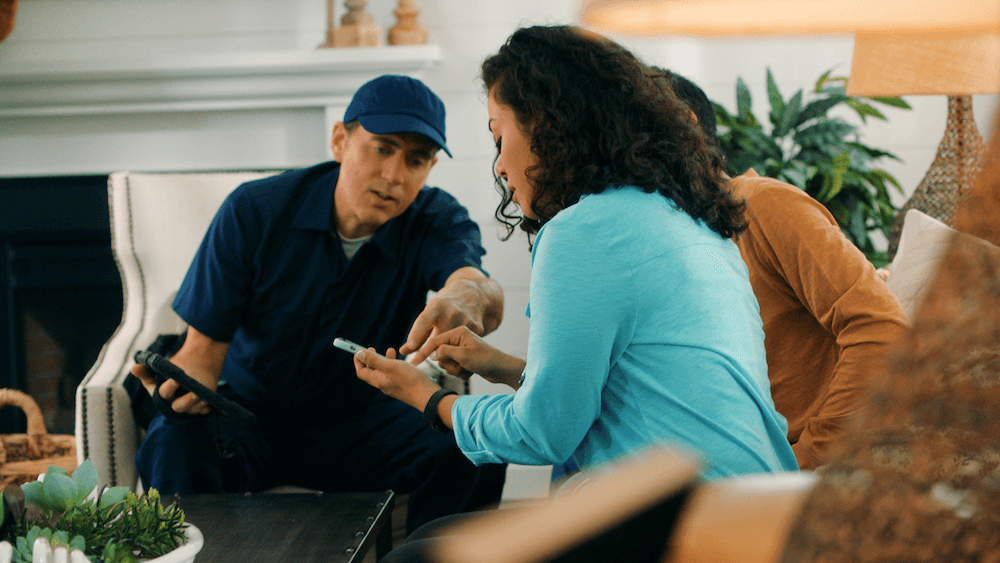 A person in a blue cap and a person in a light blue shirt are examining a smartphone together in a cozy, well-decorated living room.