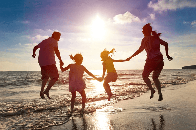 A family of four are joyfully jumping along a sunny beach. Silhouetted against a bright sun, they hold hands, evoking a sense of freedom and happiness.