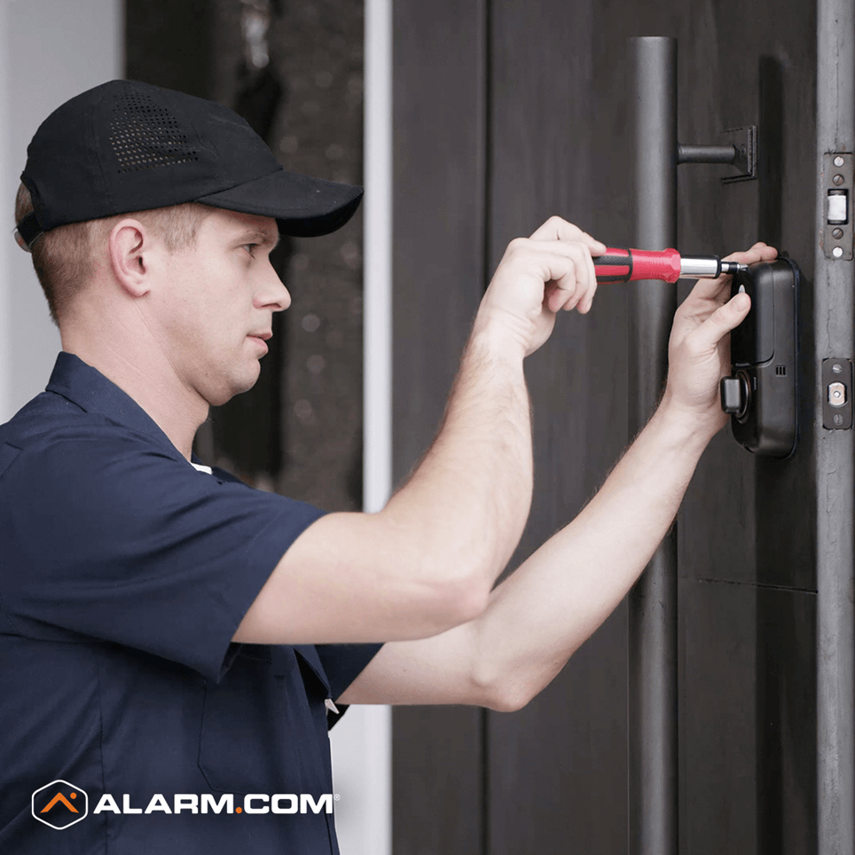 A person in a dark blue shirt and a cap is installing a smart lock on a black door using a red screwdriver, with an "ALARM.COM" logo visible.