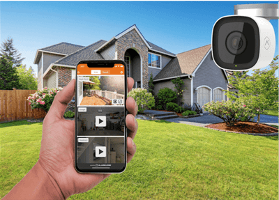 A person's hand holds a smartphone displaying security camera footage in front of a house with a lush lawn. A security camera icon floats in the corner.