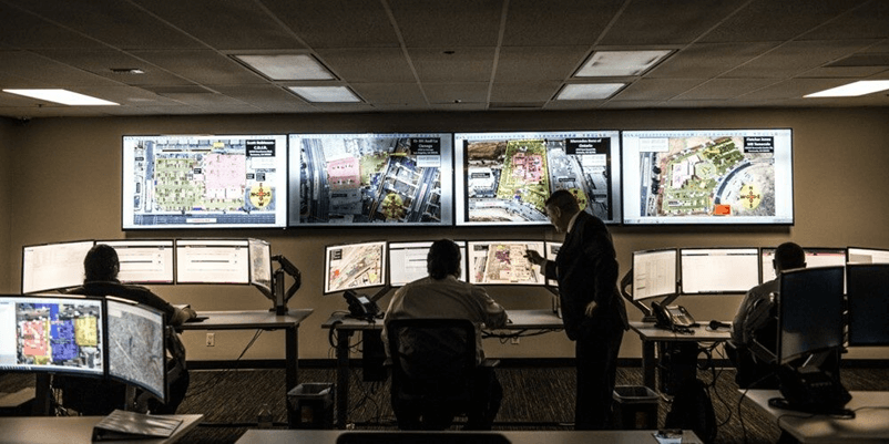 The image shows a control room with multiple people at workstations, each facing large monitors displaying maps and data. One person stands observing.