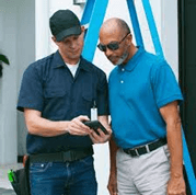 Two people are standing together; one in a cap and black shirt shows something on a handheld device to the other wearing a blue polo shirt.