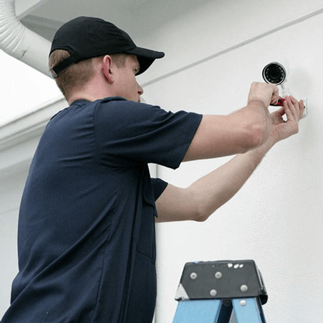 A person in a cap is installing a security camera on a white ceiling, standing on a blue stepladder. Focus on hands adjusting the device.