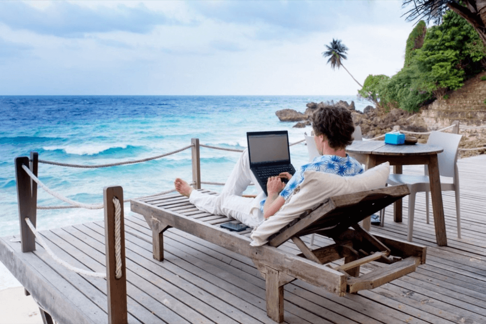 A person is working on a laptop while reclining on a wooden sun lounger overlooking a serene beach with turquoise waters, under a partly cloudy sky.