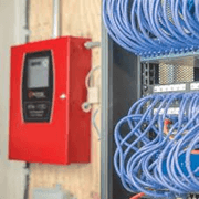 This image shows a red fire alarm pull station mounted on a wall next to a network cabinet with blue Ethernet cables, indicating a technical or server room setup.