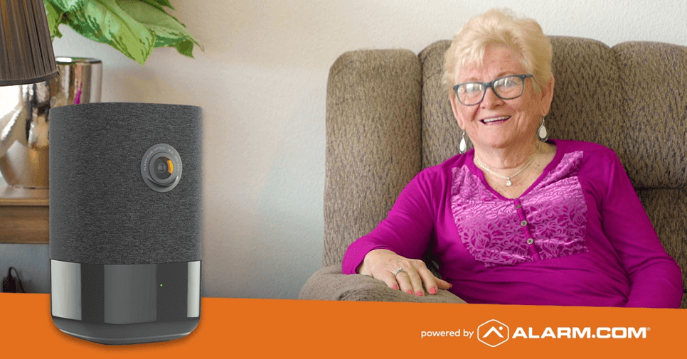 A smiling elderly person sits on a couch beside a modern smart home device with a camera, marked by the logo "powered by ALARM.COM" on an orange stripe.